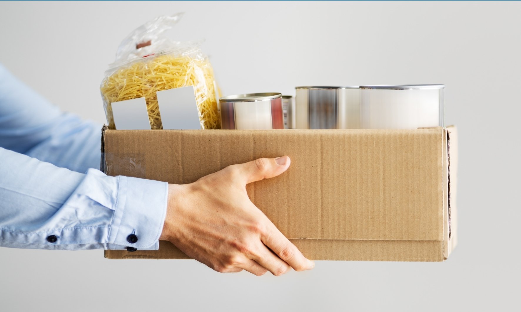 Zwei Hände, die eine Kiste mit Lebensmitteln wie Nudeln und co. tragen.